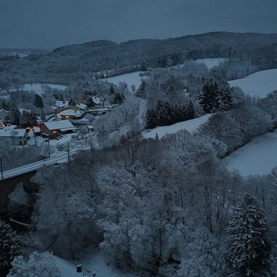 Eine Luftaufnahme einer verschneiten Landschaft mit Häusern, einer Brücke und einer kurvenreichen Straße. Die Berge sind schneebedeckt und die Bäume weiß bedeckt.
