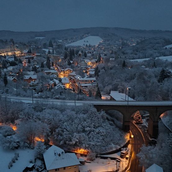 Auf einem Hügel liegt ein Dorf unter einer Schneedecke, beleuchtet vom sanften Schein der Straßenlaternen. Eine Brücke überspannt das verschneite Tal darunter.
