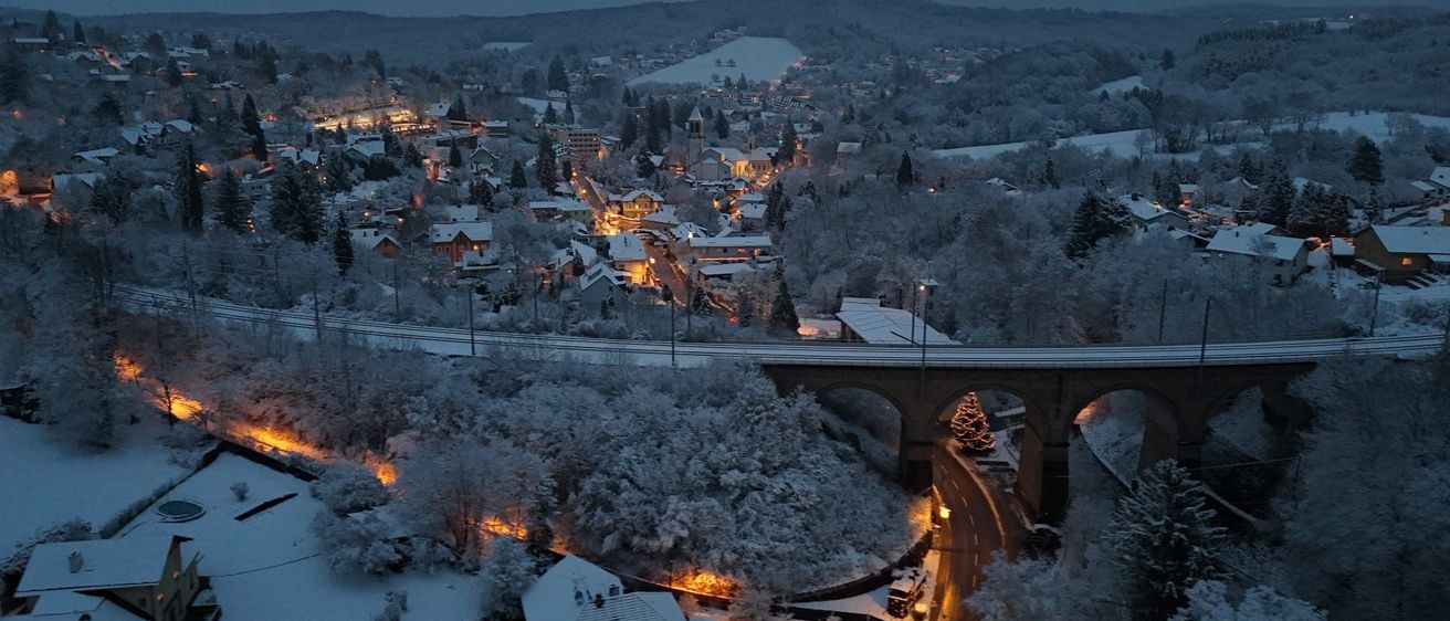 Ein Winternachtblick auf eine Stadt mit einer Brücke und einem Weihnachtsbaum darunter. Die Stadt ist schneebedeckt und es gibt Lichter.