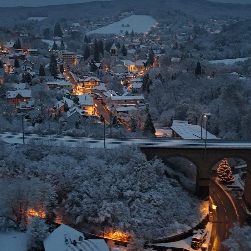 Ein Winternachtblick auf eine Stadt mit einer Brücke und einem Weihnachtsbaum darunter. Die Stadt ist schneebedeckt und es gibt Lichter.