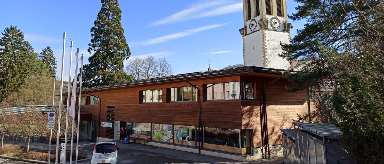 Ein modernes Gebäude mit einem Glockenturm im Hintergrund, mit Glasfenstern und einem weißen Auto davor. Bäume und Straßenlaternen sind sichtbar.