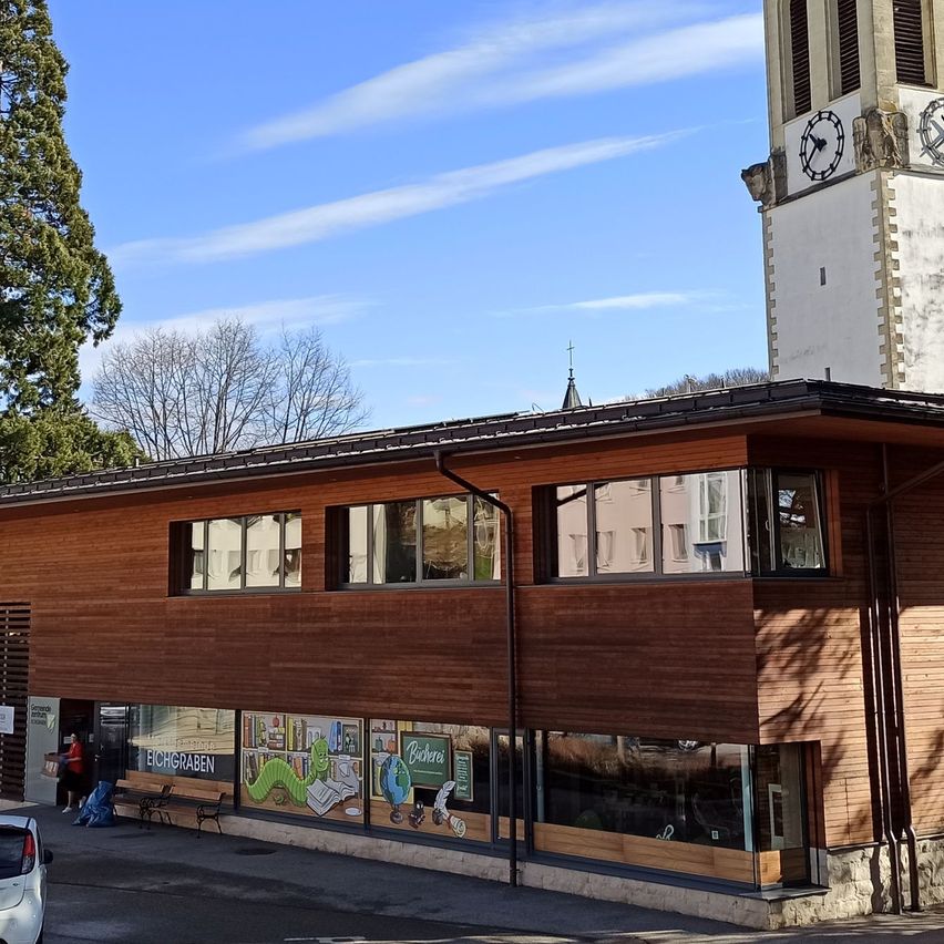 Ein modernes Gebäude mit einem Glockenturm im Hintergrund, mit Glasfenstern und einem weißen Auto davor. Bäume und Straßenlaternen sind sichtbar.