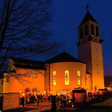 In der Nacht ist eine Kirche mit bunten Glasfenstern beleuchtet, mit einem Glockenturm und Menschen, die sich vor Ständen versammeln.