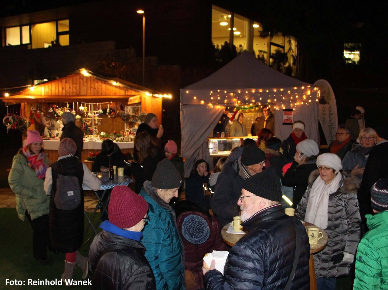 Eine Menschenmenge versammelt sich bei Nacht draußen in der Nähe von beleuchteten Ständen. Einige tragen Winterkleidung, Mützen und Brillen. Ein Mann mit weißem Bart hält ein Papier in der Hand. Ein Zelt mit Weihnachtsdekoration und Menschen darin ist sichtbar. Das Bild hat einen Wasserzeichen in der unteren linken Ecke.