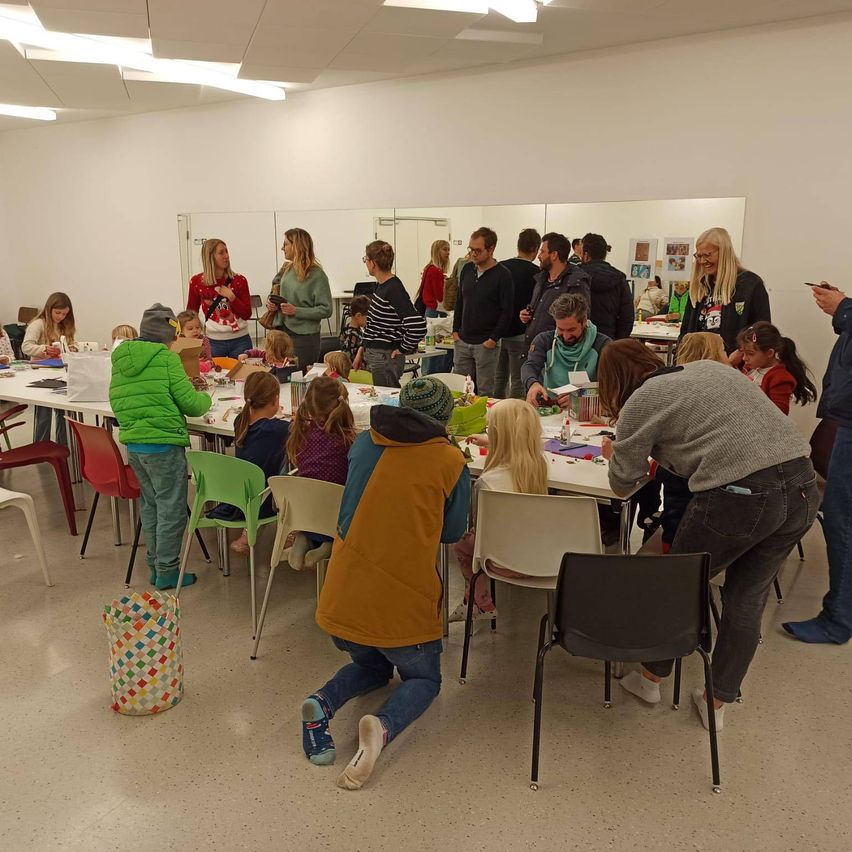 Ein Raum mit Tischen, Erwachsenen und Kindern. Sie machen wahrscheinlich eine Aktivität. Eine Frau kniet und arbeitet am Tisch.