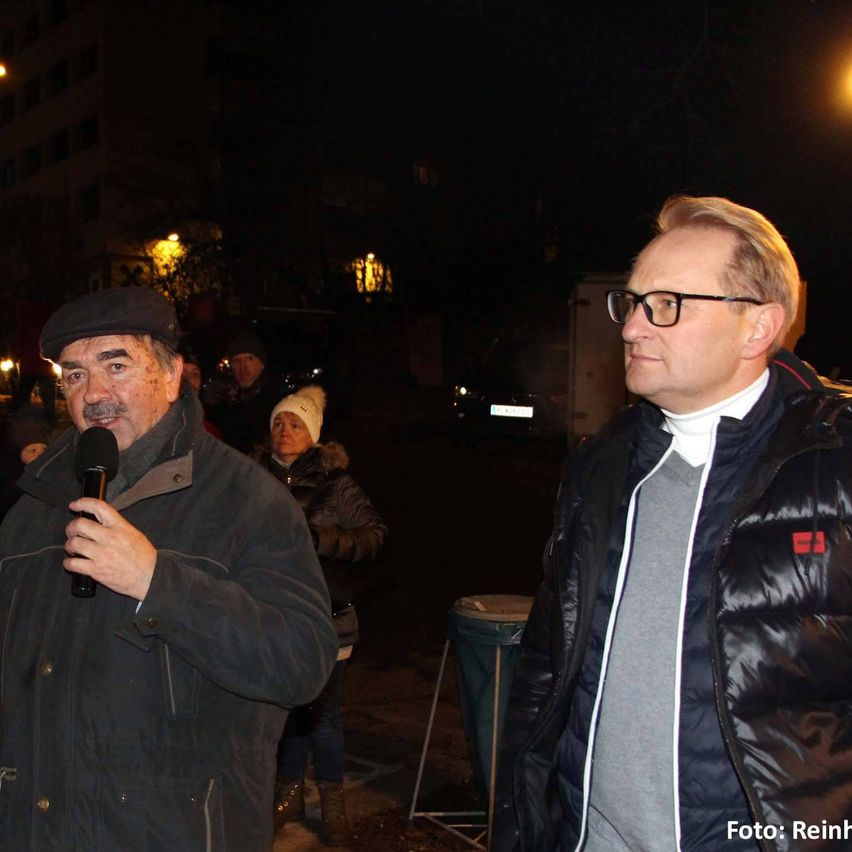 Ein Mann spricht bei Nacht im Freien in ein Mikrofon, neben ihm steht ein Mann mit Brille. Dahinter ist eine Frau mit einem fellgefütterten Hut und mehrere Personen zu sehen.