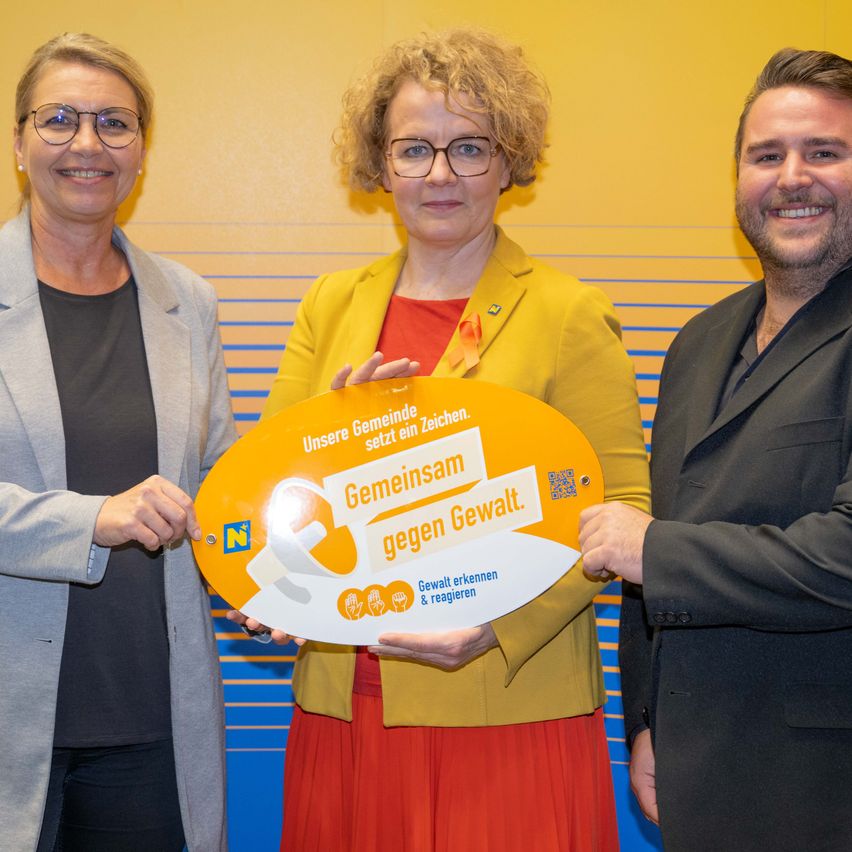 Drei Menschen stehen vor einer gelben Wand und halten ein gelbes Banner mit der Aufschrift 'Gemeinsam gegen Gewalt' in Weiß. Sie lächeln für ein Foto.