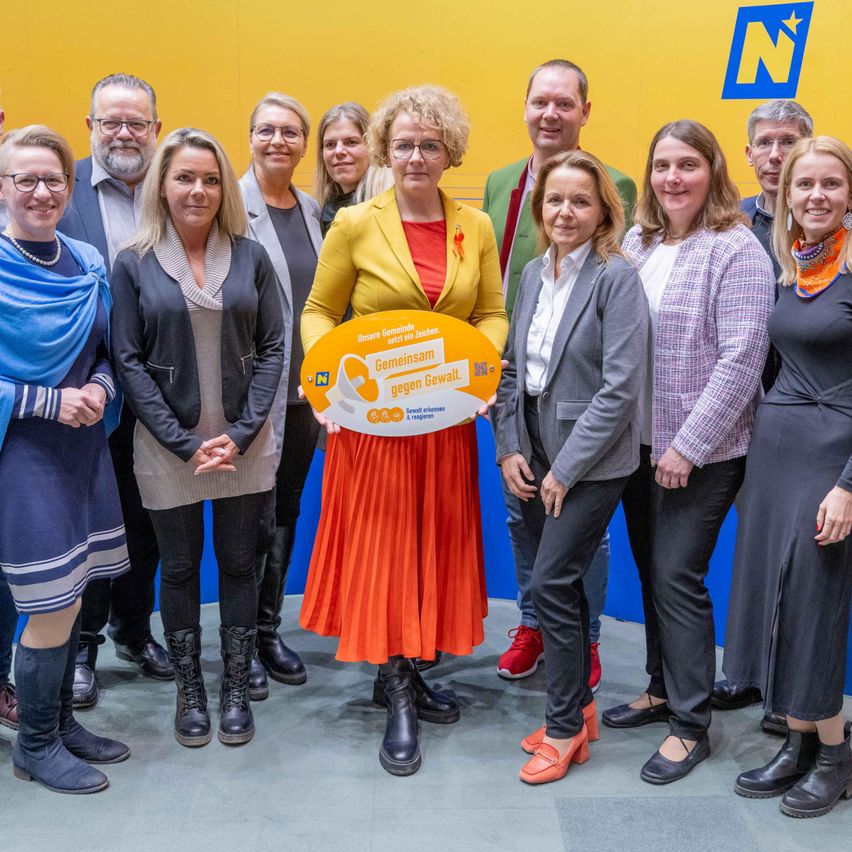 Eine Gruppe von Menschen steht lächelnd vor einer gelben Wand. Eine Frau in der Mitte hält ein Schild mit der Aufschrift 'Gemeinsam gegen Gewalt.' Dahinter ist ein blaues Logo zu sehen.