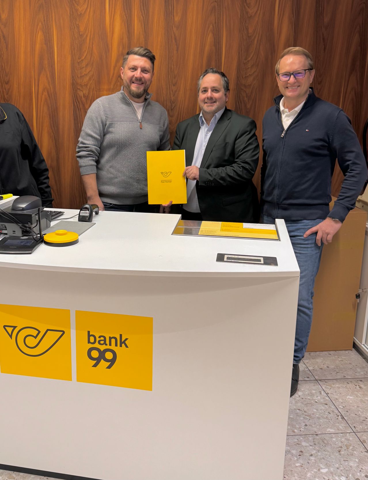 Vier Männer stehen lächelnd hinter einem Tresen und halten ein gelbes Papier. Der Tresen hat ein Bank-99-Logo. Dahinter befindet sich eine Holzwand mit einer Tür.