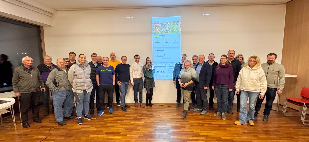 Eine Gruppe von Menschen, darunter Männer und Frauen, steht in einem Raum mit Holzboden. Einige tragen Brillen, Turnschuhe und Lederschuhe. Ein Mann in einem gelben Shirt hält einen Becher. Ein großes Poster steht hinter ihnen.