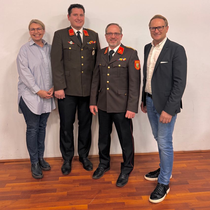 Vier Personen stehen in einem Raum mit einer weißen Wand. Drei Männer tragen Militäruniformen und eine Frau trägt eine Brille und ein Langarmhemd. Sie alle lächeln.