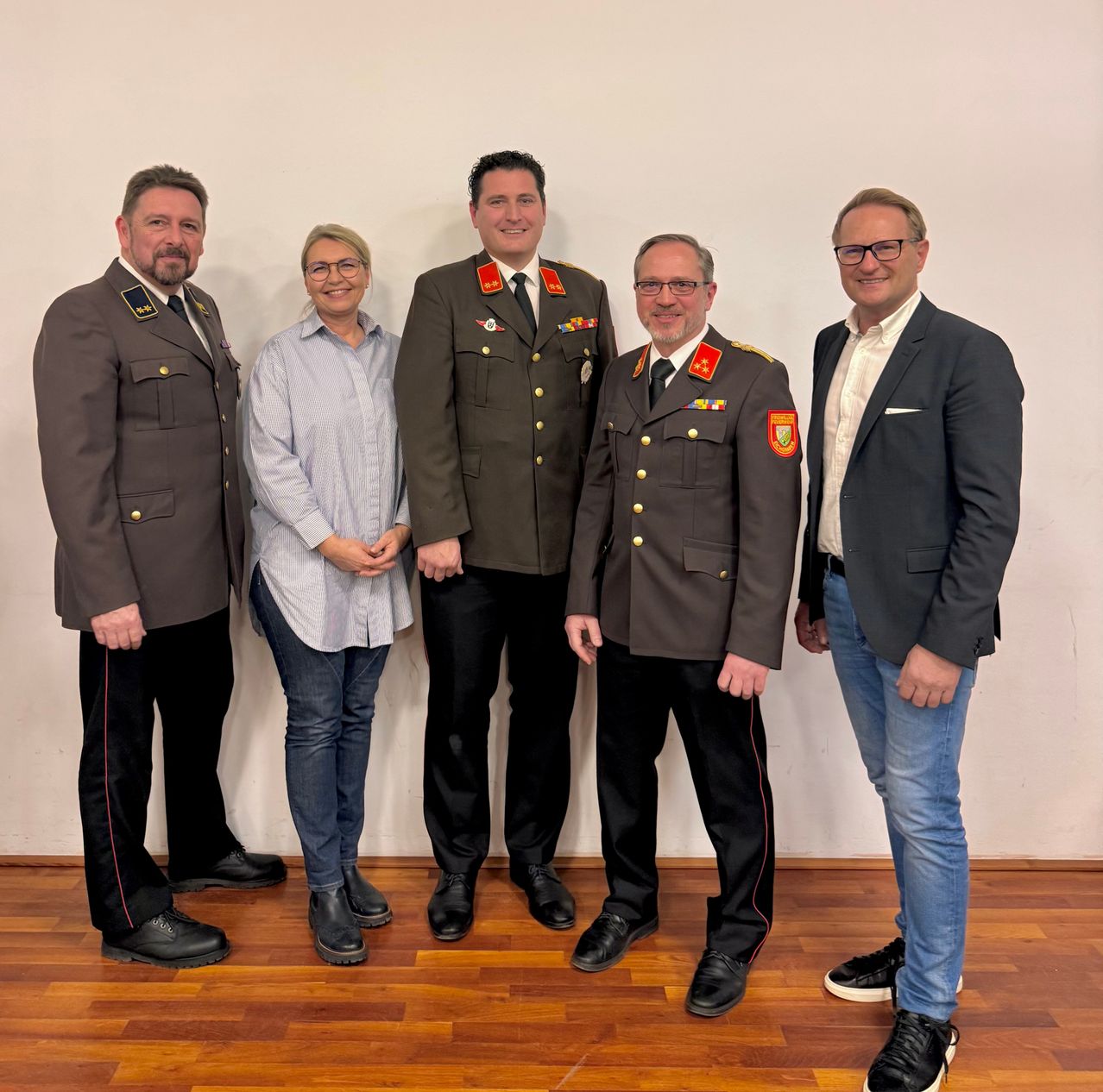 Fünf Personen, drei Männer in Uniform und zwei Männer in Anzügen, stehen zusammen vor einer weißen Wand. Sie alle scheinen für ein Foto zu lächeln.