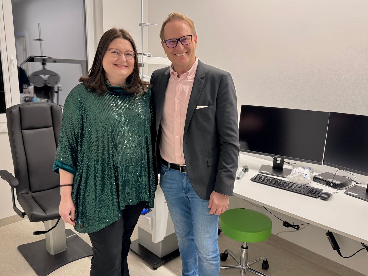 Ein Mann und eine Frau stehen in einem Raum mit einem weißen Schreibtisch und Computer. Die Frau trägt eine grüne Paillettenbluse und der Mann ein rosa Hemd und einen grauen Anzugsakko. Sie lächeln für die Kamera.