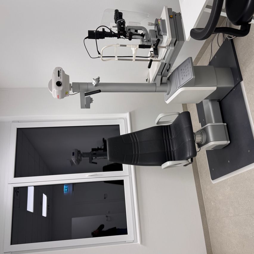 Ein Untersuchungsraum mit einem ophthalmologischen Diagnosegerät auf einem Ständer, begleitet von einem schwarzen Liegestuhl. Der Raum ist gut beleuchtet, wobei natürliches Licht durch ein großes Fenster eintritt.