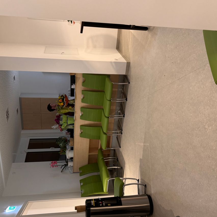 Eine Draufsicht eines Raums mit einem Holztisch, sechs grünen Stühlen und einer Person, die Blumen arrangiert. Der Raum hat weiße Wände, geflieste Böden und ein Fenster mit Blick nach draußen.