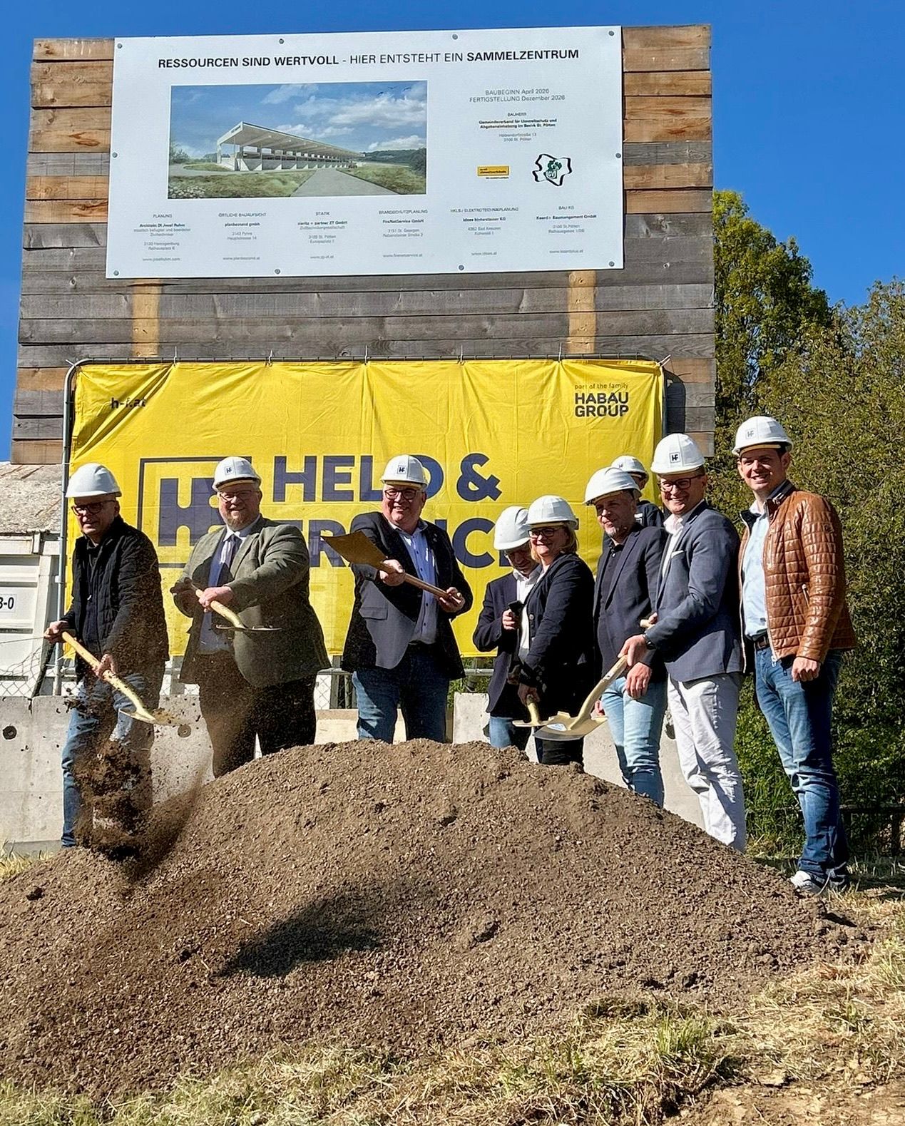 A group of people in hard hats are at a construction site, posing for a photo while holding shovels. Behind them, a wooden wall with a yellow banner displays the words Helbig & Company.
