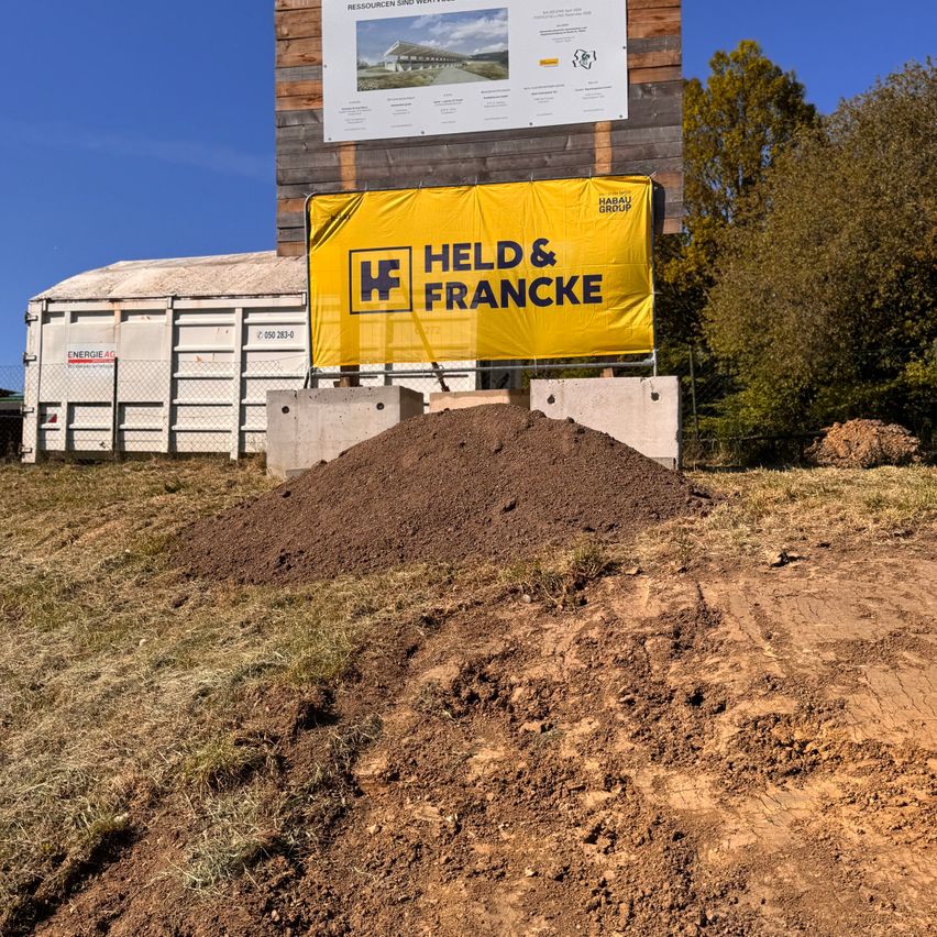 A construction site with a large pile of dirt and a yellow banner reading 'HELD & FRANCK' with an image of a building. A wooden billboard is behind it, and there are trees in the background.