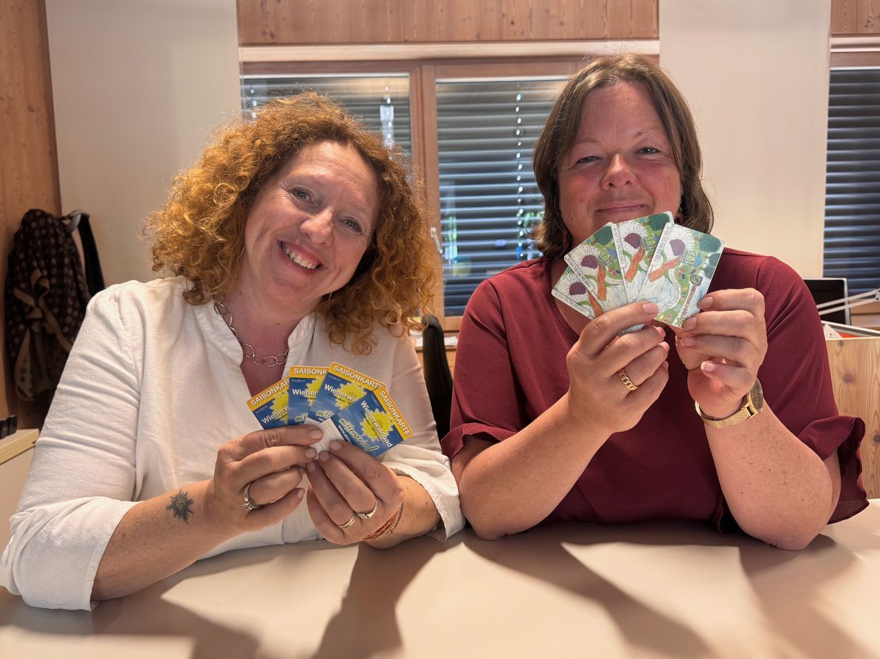 Two women smiling and holding up cards, sitting at a table in a room with a window and wooden wall.
