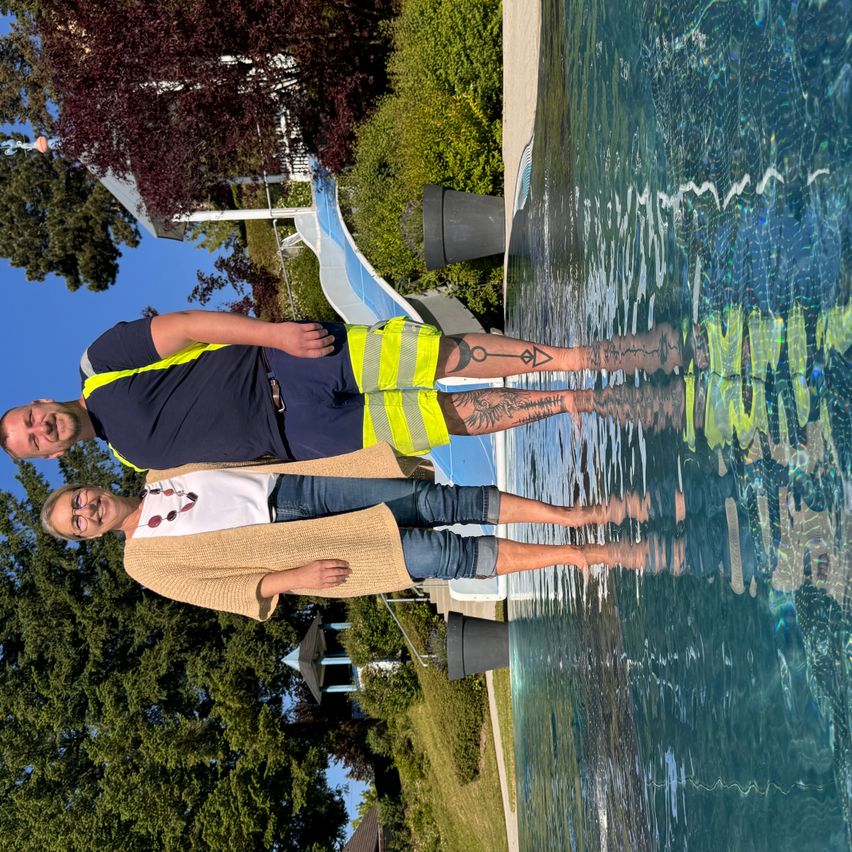 Zwei Personen stehen in einem Pool, einer trägt eine gelbe Warnweste und der andere eine braune Strickjacke. Sie lächeln beide in die Kamera. Hinter ihnen befindet sich eine Wasserrutsche und einige Bäume.