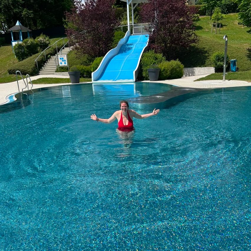 Eine Frau in einem roten Badeanzug steht in einem großen Schwimmbecken, Arme ausgestreckt. Hinter ihr befindet sich eine blaue Wasserrutsche.
