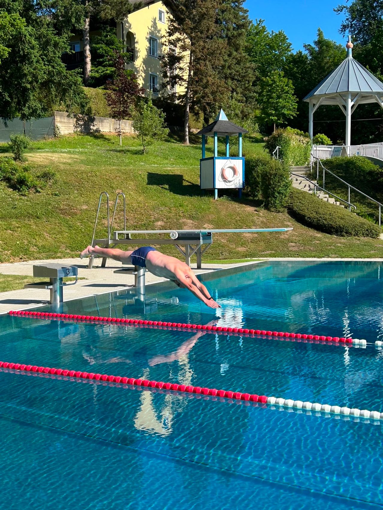 Eine Person taucht in ein Schwimmbecken mit roten Bahnmarkierungen. Das Becken ist von einer Rasenfläche und einer Sprungturm-Plattform umgeben.