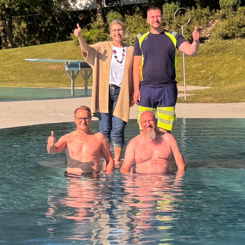 Drei Personen stehen in einem Pool und lächeln. Zwei Männer geben Daumen hoch, einer in Badeshorts. Eine Frau in einem Strickjacken steht hinter ihnen. Ein Tischtennistisch und ein Zaun sind im Hintergrund zu sehen.