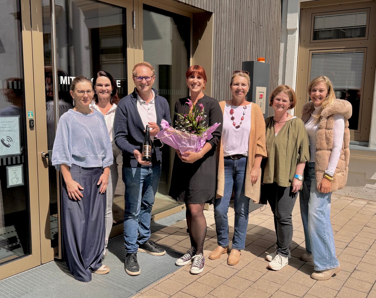 Eine Gruppe von Menschen steht vor einem Gebäude. Eine Frau in der Mitte hält Blumen und eine Flasche. Der Mann neben ihr hält ebenfalls eine Flasche. Sie alle lächeln.