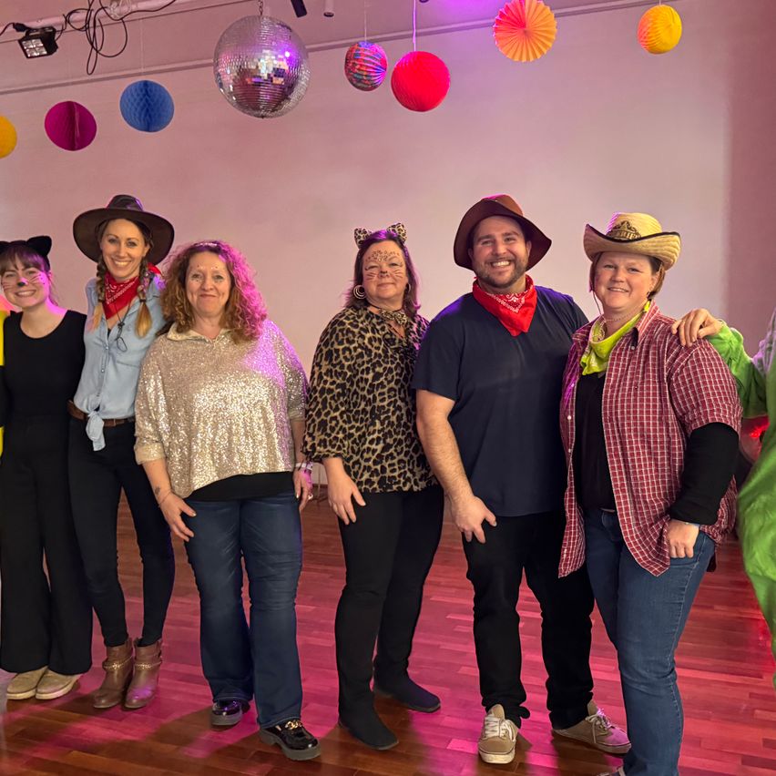 A group of people dressed in costumes pose for a photo in a room with colorful balloons and a disco ball.