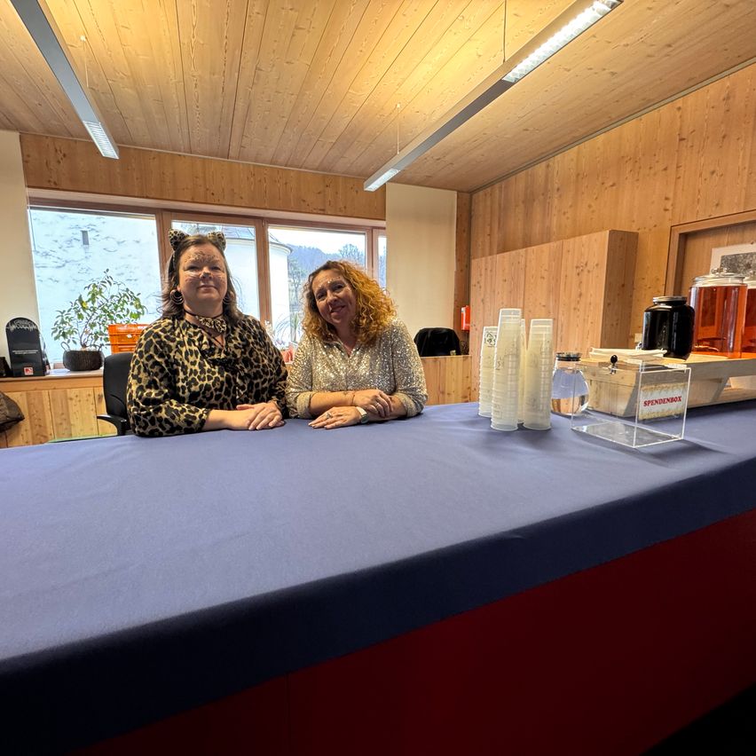 Two women are smiling while sitting at a long table. They have cups, a water pitcher, and a box in front of them. Behind them is a wooden wall with windows and a potted plant.