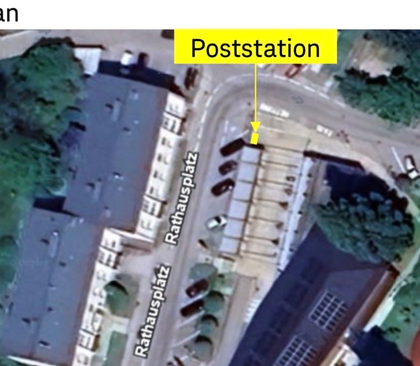 Eine Luftaufnahme zeigt eine Stadtstraße mit mehreren Gebäuden. Ein gelbes Schild zeigt 'Poststation' und ein gelber Pfeil weist auf einen bestimmten Ort an der Straße. Mehrere Autos sind auf der Straße geparkt.