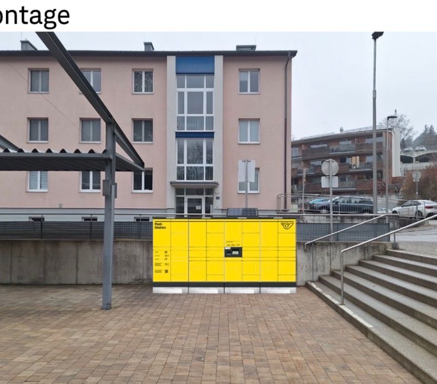 Ein gelber Poststation-Paketautomat ist in einem gepflasterten Außenbereich installiert, mit Treppen, die zu einem Geländer führen. Ein Gebäude mit mehreren Fenstern befindet sich hinter dem Automaten.