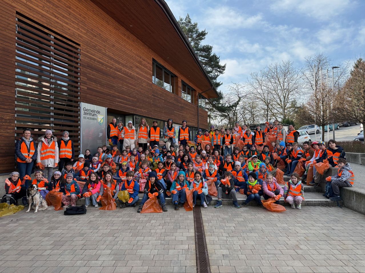 Eine Gruppe von Kindern und Erwachsenen in orangenen Westen posiert für ein Foto vor einem Gemeindezentrum und hält Mülltüten in den Händen.