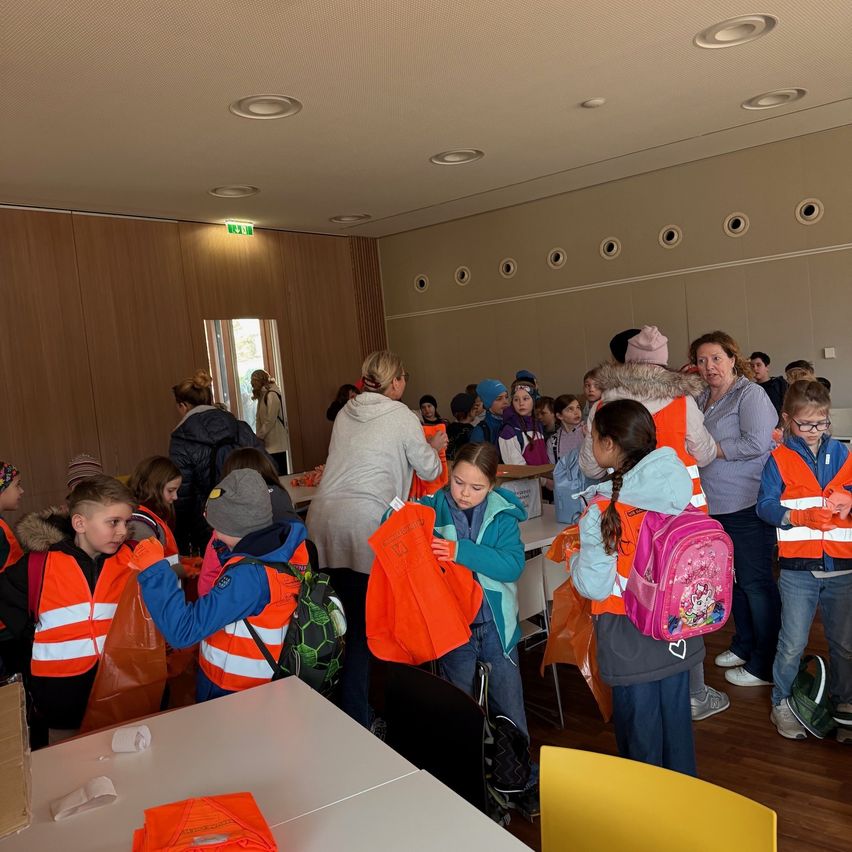 Eine Gruppe von Kindern und Erwachsenen versammelt sich in einem Raum, alle tragen orange Sicherheitswesten. Sie stehen um einen Tisch mit Taschen und bereiten sich möglicherweise auf eine Veranstaltung vor.