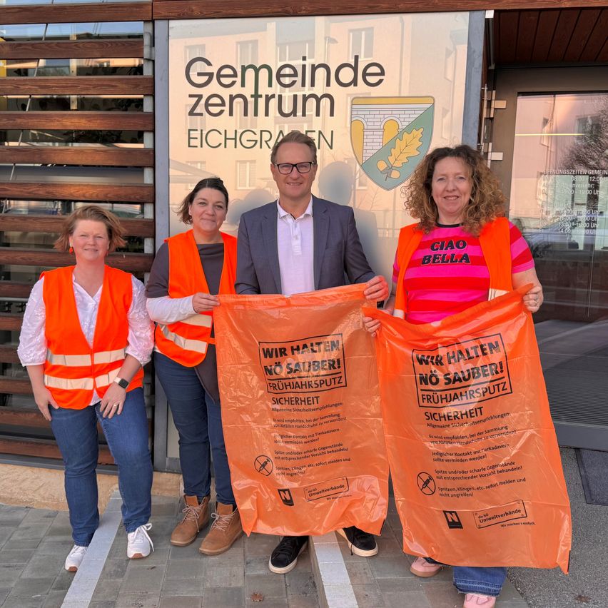 Vier Personen posieren vor einem Gebäude mit der Aufschrift Gemeinde zentrum Eichgraben und halten orangefarbene Beutel mit Sicherheitshinweisen.