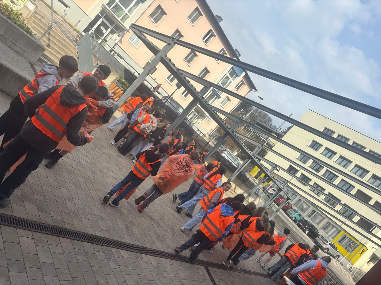 Eine Gruppe von Menschen in orangen Sicherheitswesten und Turnschuhen steht draußen in der Nähe von Gebäuden und geparkten Autos.