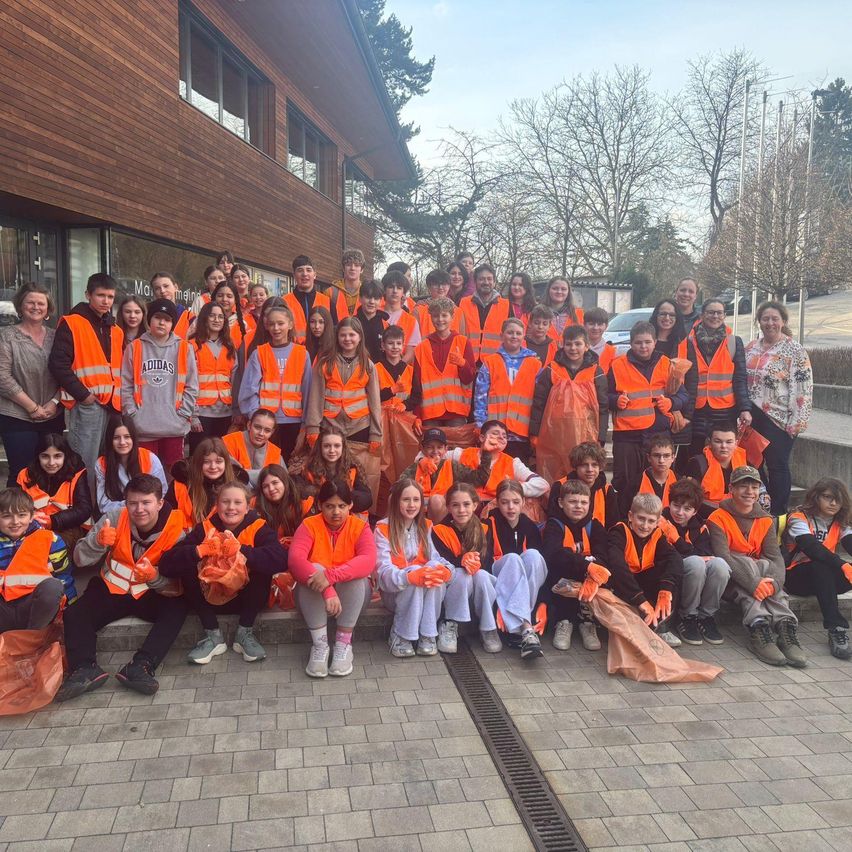 Eine Gruppe von Menschen in orangefarbenen Warnwesten posiert für ein Foto vor einem Gebäude an einem sonnigen Tag.