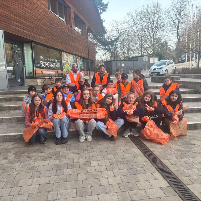 Eine Gruppe von Kindern in orangefarbenen Westen posiert für ein Foto vor einem Gebäude. Sie halten Plakate mit deutschem Text. Zwei Erwachsene, einer mit Bart, sind anwesend.