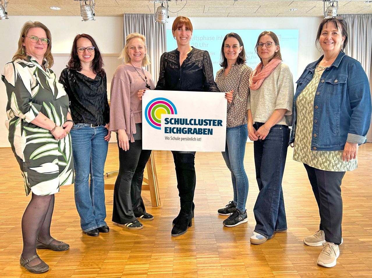 Sechs Frauen stehen in einem Raum und lächeln. Eine Frau hält ein Schild, auf dem 'Schulcluster Eichgraben' steht. Sie scheinen ein Ereignis zu feiern oder daran teilzunehmen.
