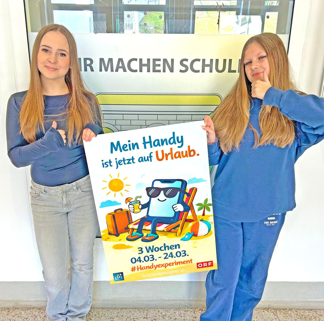 Zwei Frauen in passenden blauen Outfits stehen neben einer Glastür. Eine hält ein Schild mit einem Cartoon-Charakter, einer Strandszene und dem Text 'Mein Handy ist jetzt auf Urlaub.' Sie geben beide einen Daumen hoch.
