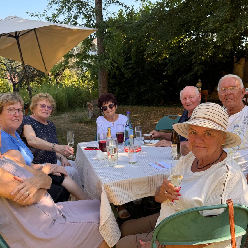 Eine Gruppe älterer Frauen und Männer sitzt um einen Tisch in einem Garten und posiert für ein Foto. Der Tisch hat Weingläser, Flaschen und eine weiße Tischdecke. Dahinter steht ein Baum und ein Haus.