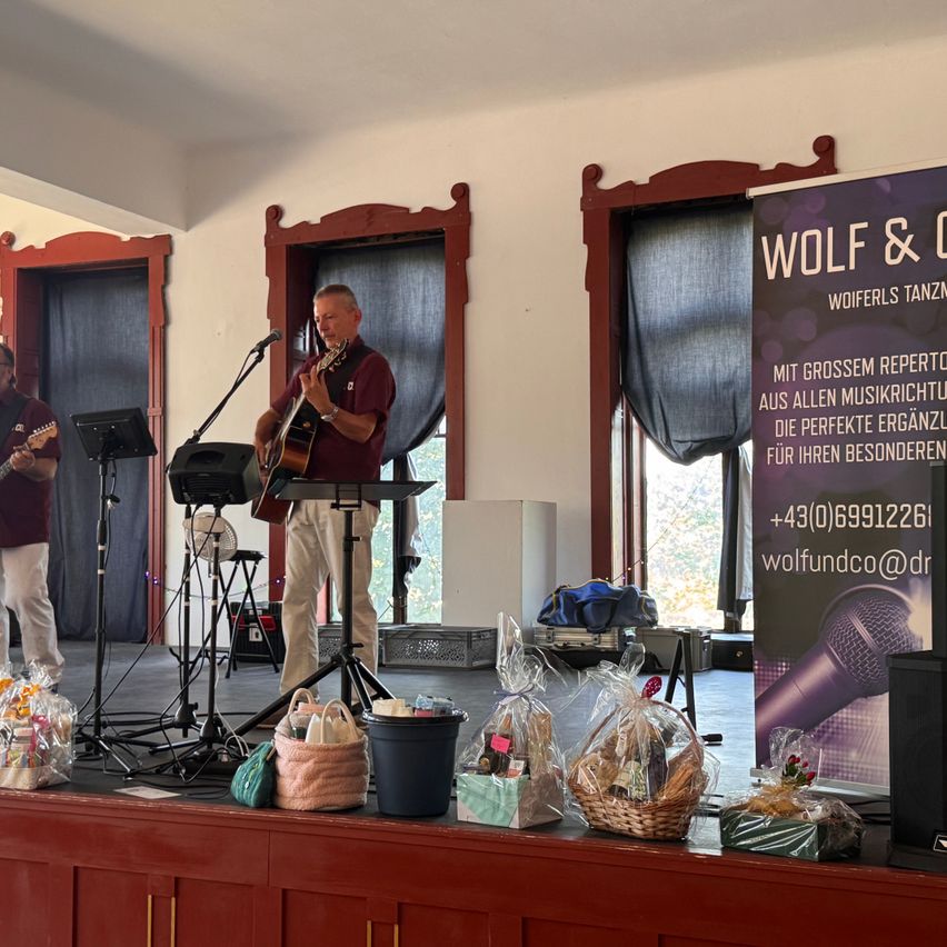 Zwei Männer spielen auf der Bühne, einer spielt Gitarre und der andere spielt Gitarre und singt. Ein Banner mit dem Text 'WOLF & CO' ist auf der rechten Seite sichtbar. Auf der Bühne befinden sich verschiedene Gegenstände, darunter ein Mikrofon, ein Lautsprecher und ein Korb.