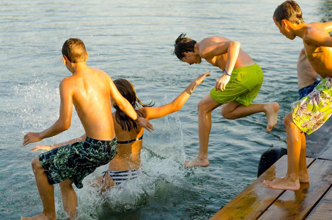 Drei Kinder spielen im Wasser, ein Junge springt hinein, ein anderer Junge streckt die Hand aus und ein Mädchen schaut sie an.