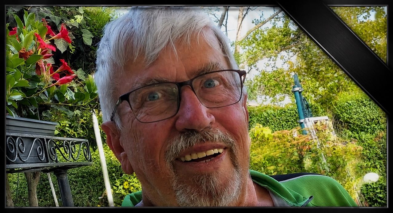 Ein älterer Mann mit weißem Haar und Bart, Brille tragend, lächelt in einem Garten mit grünen Büschen und Bäumen im Hintergrund.