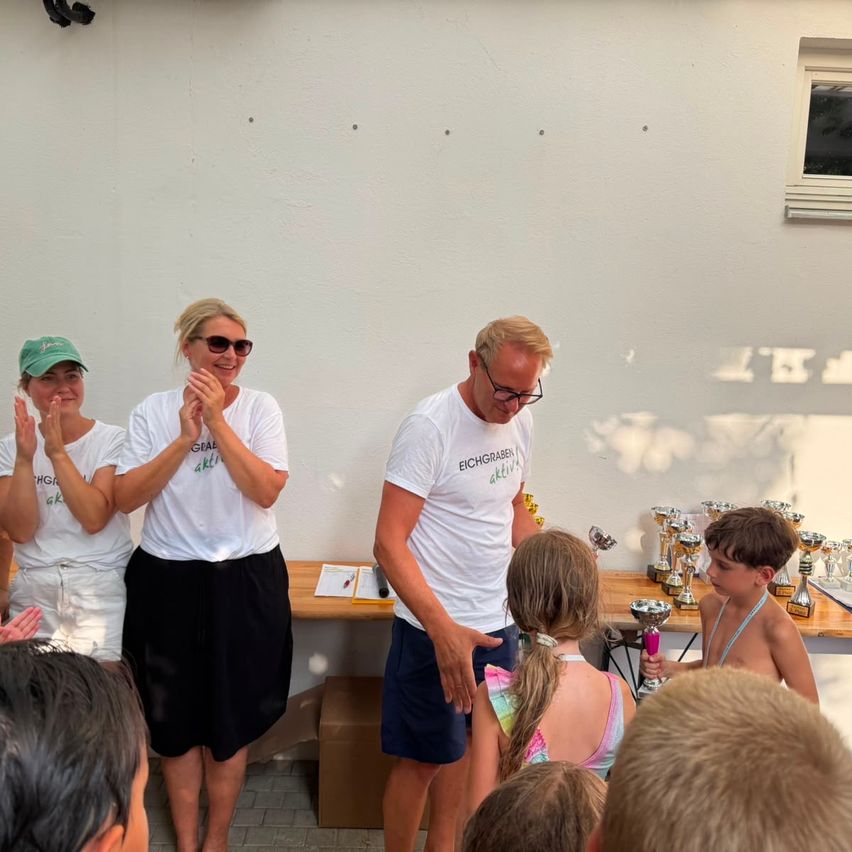 Eine Gruppe von Menschen, darunter ein Mann und zwei Frauen, steht vor einer Wand mit einem Pokal auf einem Tisch. Der Mann hält ein junges Mädchen, und die beiden Frauen klatschen. Das Mädchen hält einen Pokal, und ein Junge steht neben ihr. Auf dem Tisch befinden sich mehrere Pokale.