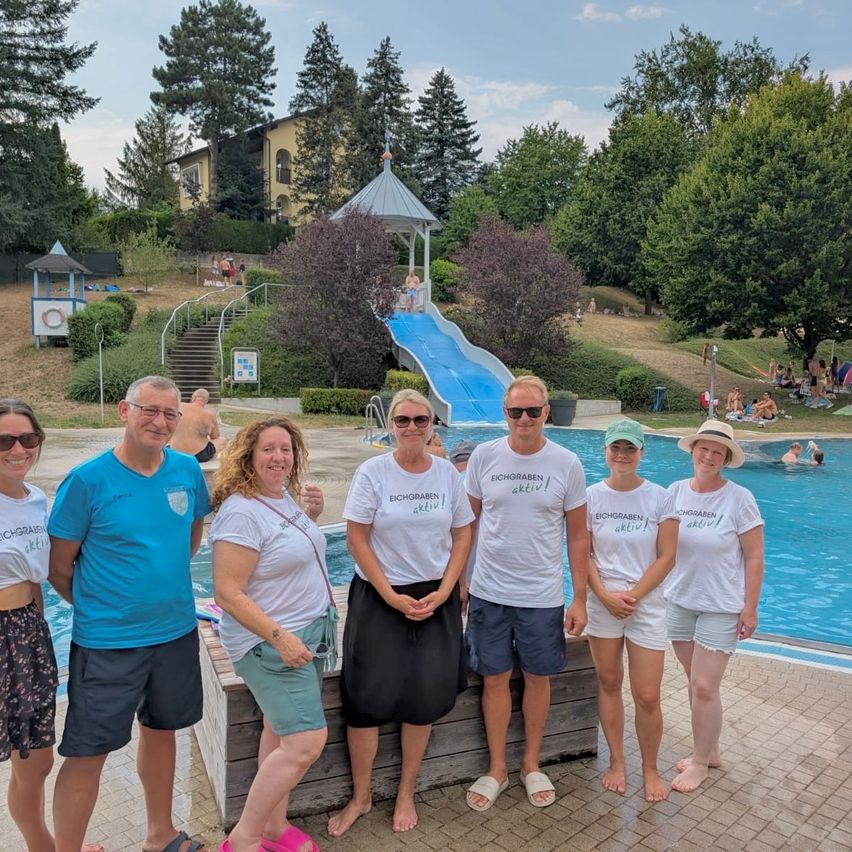 Eine Gruppe von Menschen in passenden weißen T-Shirts mit Eichgraben aktiv darauf, stehen vor einem Pool. Dahinter ist eine blaue Wasserrutsche und ein Gebäude zu sehen.