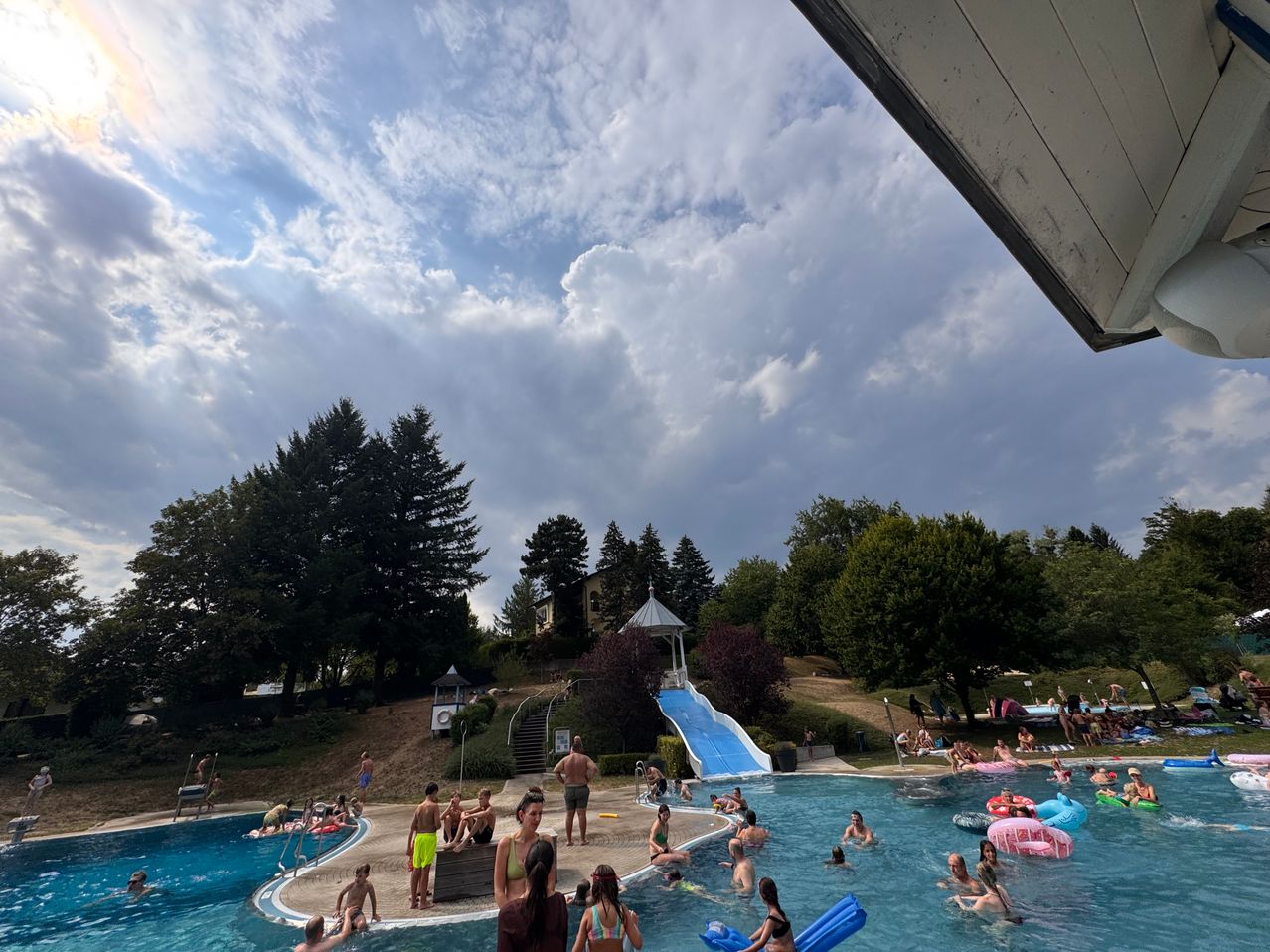 Eine Gruppe von Menschen genießt einen Tag im Freibad, einige schwimmen, während andere auf aufblasbaren Schwimmhilfen entspannen, unter einem teilweise bewölkten Himmel.