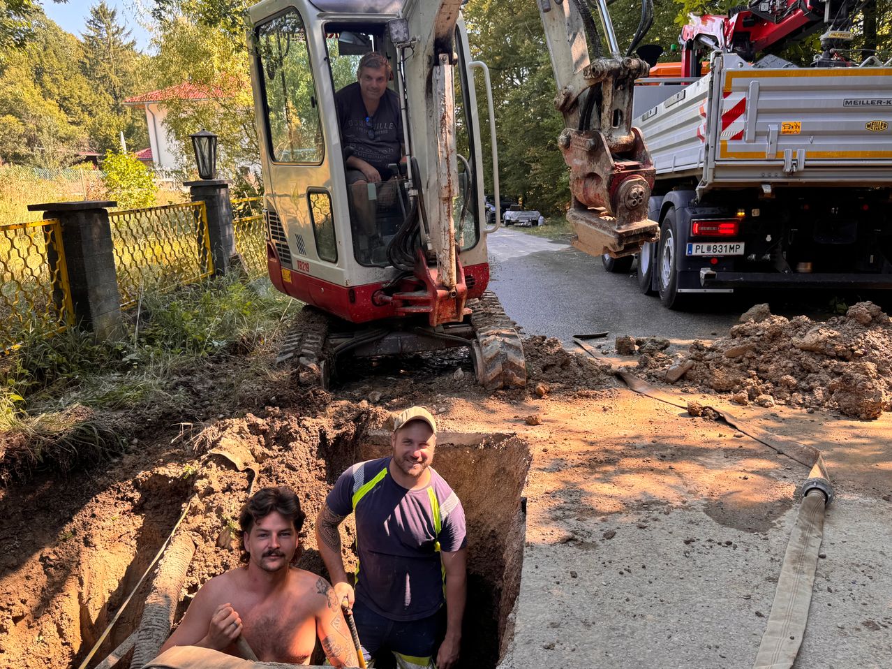 Zwei Männer stehen in einem Loch mit einem großen Bagger dahinter. Ein Mann ist ohne Hemd und hält eine Schaufel, während der andere eine Mütze und eine Warnweste trägt. Ein Lkw ist am Straßenrand geparkt.