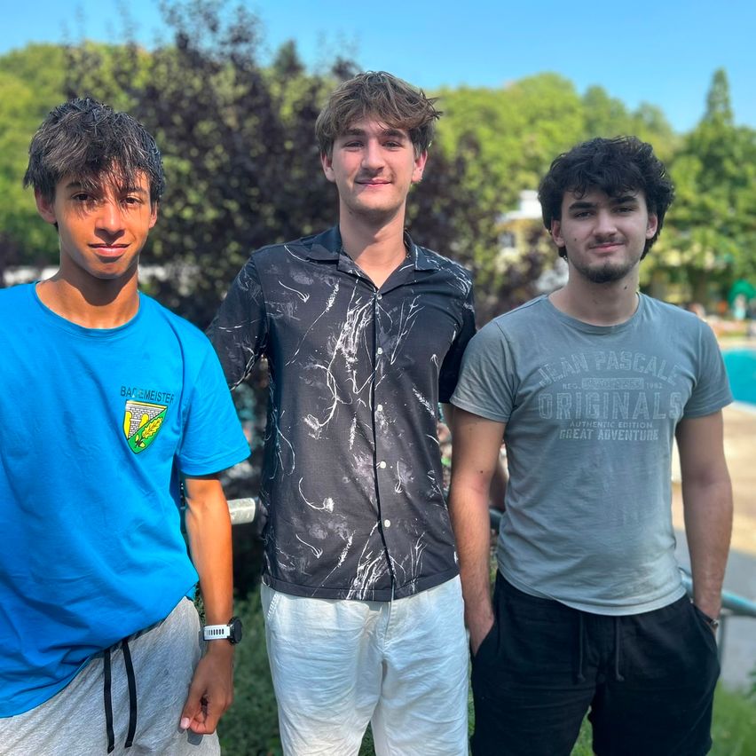 Three young men stand together in a park, smiling for the camera. The man on the left wears a blue shirt with a logo, the middle man a black shirt with a design, and the man on the right a gray shirt with text.