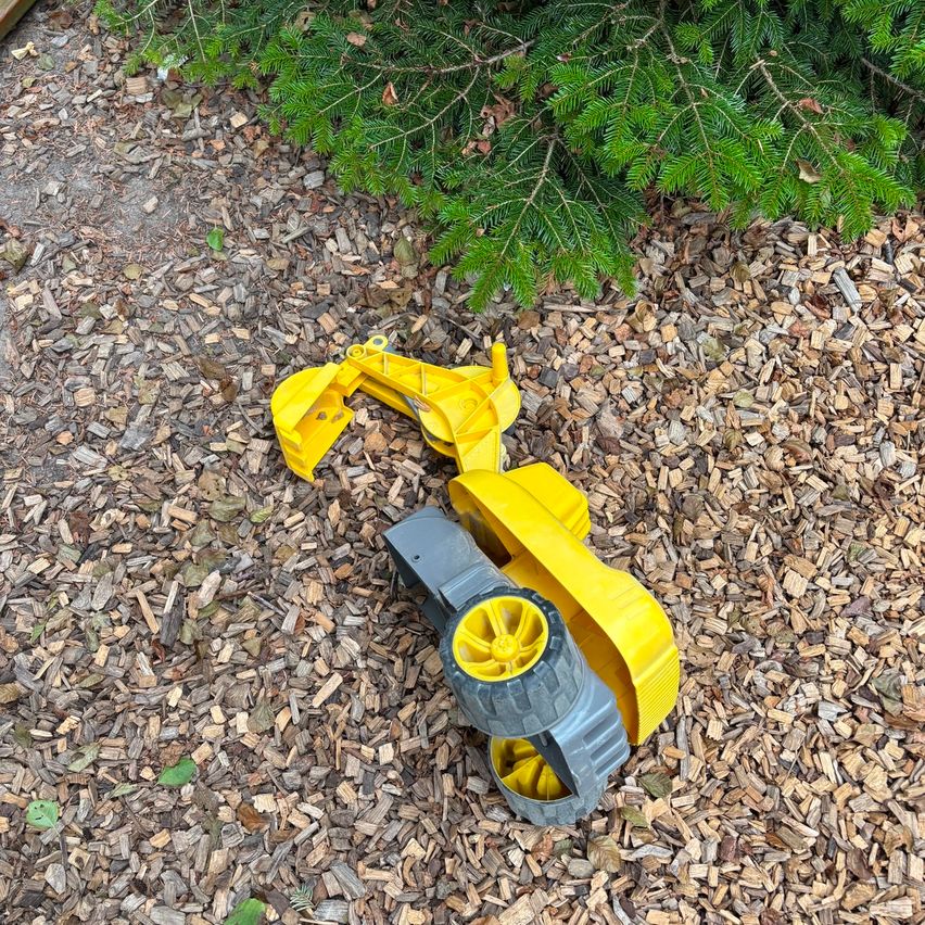 Ein gelber und grauer Spielzeugbagger liegt auf dem mit Holzspänen bedeckten Boden.