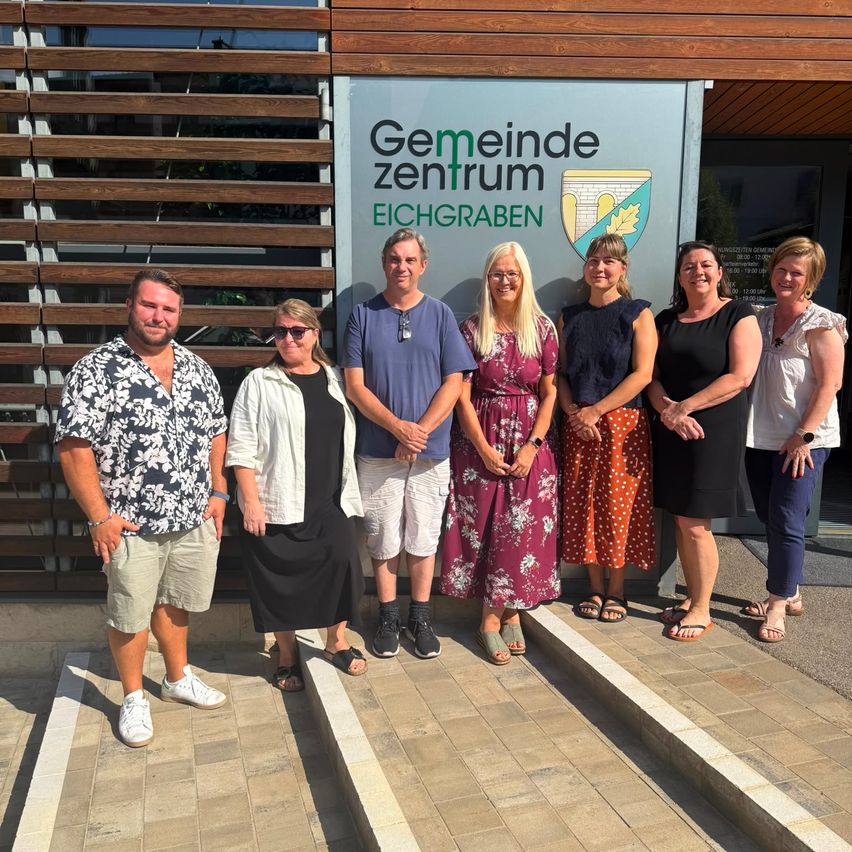 A group of people stands in front of a building with the sign 'Gemeinde zentrum Eichgraben'. They are all smiling and appear to be posing for a photo.